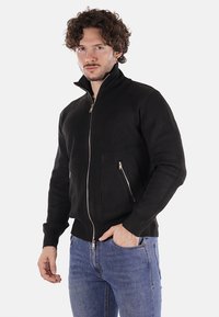 Giacca nera a coste con zip e colletto alto, dotata di due tasche laterali e un design aderente. Tessuto testurizzato.