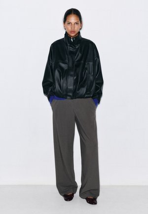 Personne portant une veste en cuir noire, un pantalon gris à jambes larges et des chaussures foncées, debout contre un mur blanc uni, les mains dans les poches.