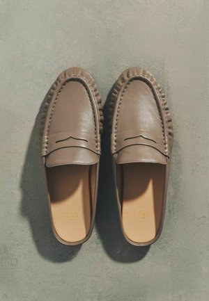 Par af brune læder slip-on loafers med syningsdetaljer, vist oppefra på en grå tekstureret overflade.