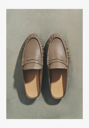 Pár hnedých kožených mokasín bez šnúrok s prešitým zdobením, zobrazených zhora na sivom textúrovanom povrchu.