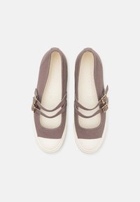 Zapatos de lona marrones tipo slip-on con dos correas diagonales y hebillas en tono dorado. Punta de goma blanca y plantilla texturizada. Diseño plano.