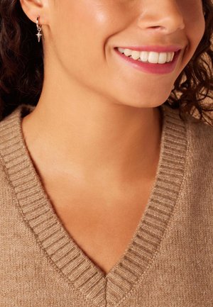 Hängende Creolen mit einem spitzen Anhänger, aus silberfarbenem Metall, getragen von einer Person mit dunklem, lockigem Haar. V-Ausschnitt, beiger Strickpullover sichtbar.