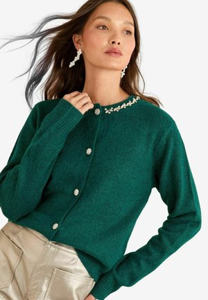 Grüne Strickjacke mit runder Ausschnittform, dekorativen Strassakzenten am Kragen und perlenähnlichen Knöpfen. Der Stoff wirkt weich und strukturiert.