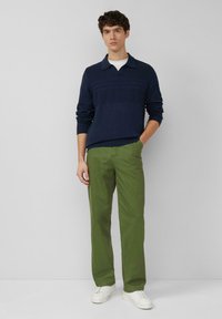 Jonge man die met één hand in zijn zak staat, draag een marineblauwe trui, olijfgroene broek en witte sneakers tegen een effen achtergrond.