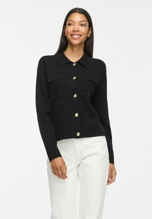Femme aux longs cheveux foncés porte un cardigan noir boutonné avec des boutons dorés et un pantalon blanc, souriante et posant devant un fond blanc.