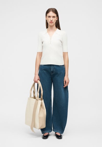 Γυναίκα με ίσια καστανά μαλλιά, φορώντας λευκό πλεκτό τοπ με κοντά μανίκια και μπλε τζιν παντελόνι με φαρδιά πόδια, κρατώντας μια μεγάλη μπεζ τσάντα ώμου στο αριστερό χέρι.