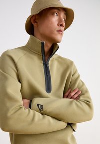 Χακί φλις πόλο με ψηλό γιακά και μαύρο φερμουάρ, διαθέτοντας το μαύρο λογότυπο Nike στο μανίκι. Λείος υφή και εφαρμοστό σχέδιο.