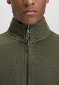 Olive green zip-up jacket made of soft fabric, featuring a high collar and a front zipper. Smooth texture with minimal stitching details.