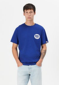 T-shirt in cotone blu con maniche corte, scollatura rotonda e un logo circolare bianco che presenta '981' e 'REFLAY' sul petto.