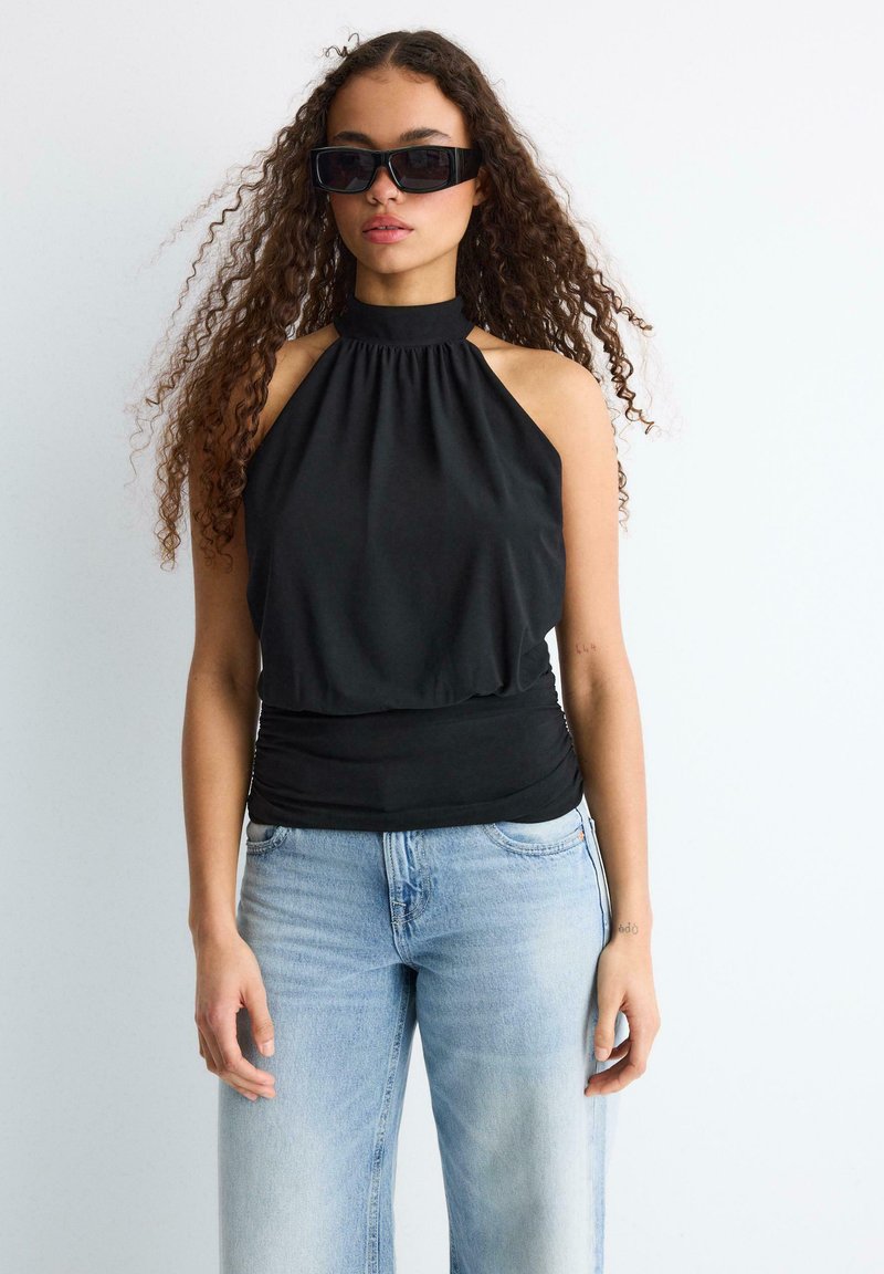 Jeune femme aux cheveux bouclés portant un top noir sans manches à col halter, un jean bleu clair et de grandes lunettes de soleil noires sur fond blanc.