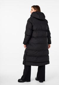Doudoune noire avec capuche, design long, texture matelassée, boutons pression sur le côté, et finition élégante, portée sur un pantalon noir et des bottes.