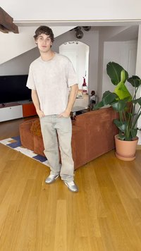 Világosszürke, oversize t-shirt, világoskék farmernadrág és ezüstszínű sportcipők. A háttérben csíkozott szőnyeg és egy barna kordbársony kanapé található, mellette egy növény.