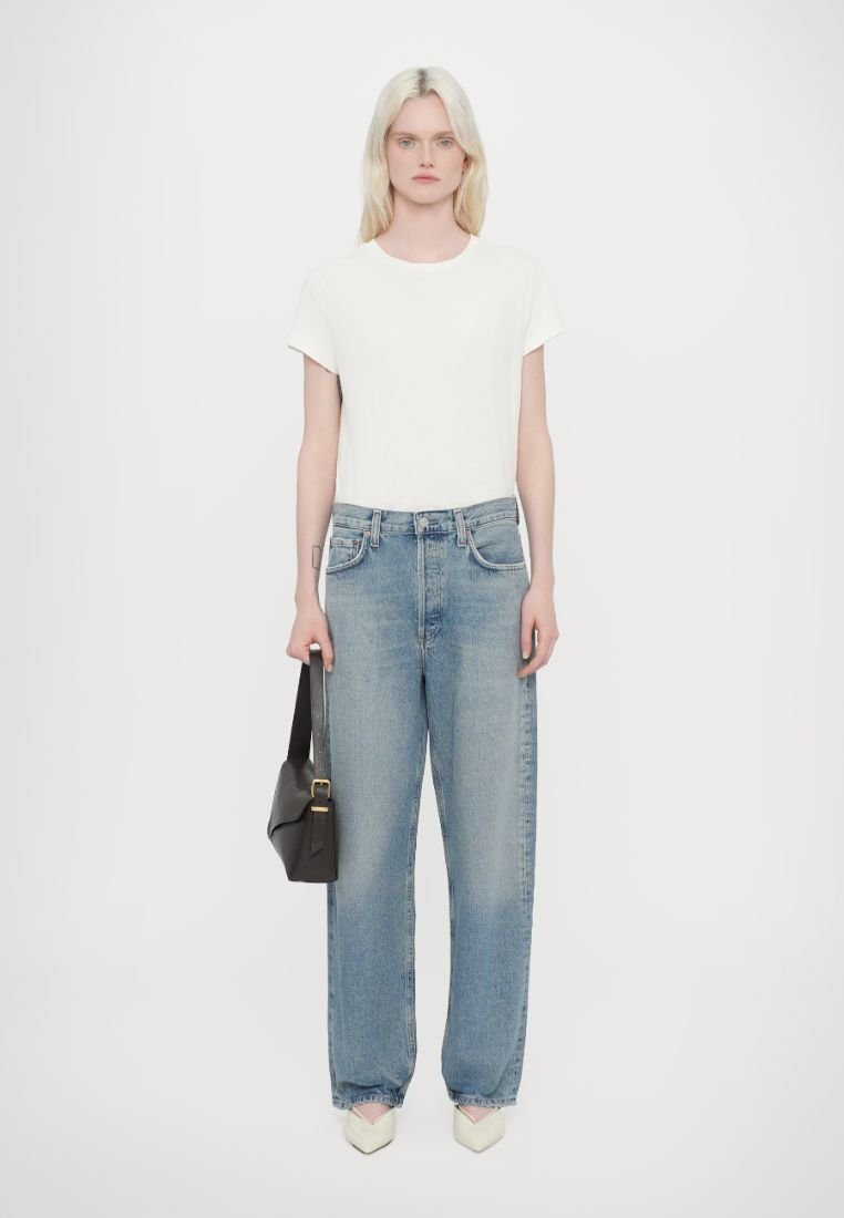 T-shirt blanc à manches courtes, jean droit taille haute bleu clair, ballerines beige à bouts pointus et sac à bandoulière noir. Fond uni.