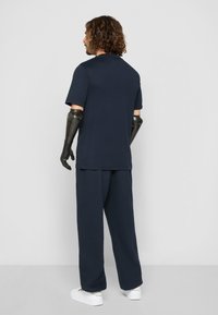 Nachtblaues Baumwoll-T-Shirt und passende Hose. Mit kurzen Ärmeln, lockerer Passform und weißen Sneakern. Strukturierte schwarze Prothesenarme.