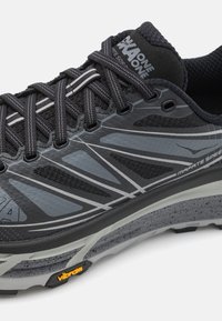 Chaussure de course noire avec des accents gris, tige en mesh et semelle en caoutchouc texturé. Présente des lacets visibles, des bandes réfléchissantes et la marque Vibram.