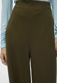 Höga, mörk olivgröna byxor i mjukt tyg med subtila frontveck, kombinerade med en ljusblå långärmad topp.