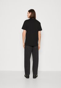 Zwarte T-shirt met korte mouwen gezien vanaf de achterkant, gecombineerd met donkergrijze broeken. Eenvoudig, onopgesmukt ontwerp met een ontspannen pasvorm.