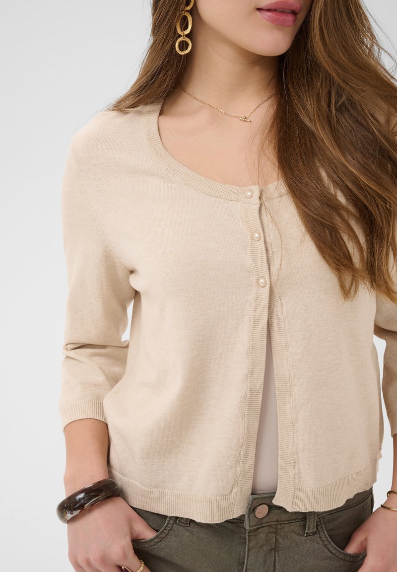 Femme portant un cardigan beige boutonné sur un haut clair, un pantalon olive, des boucles d'oreilles créoles en or, un collier délicat et un bracelet foncé.