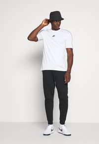 Vit Nike t-shirt med svart logotyp, svarta joggers med paneldetaljer och en svart bucket hat, kompletterat med vita Nike-skor.