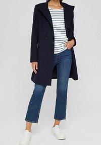 Marineblauwe dubbelzijdige jas met grote revers, gedragen over een wit-blauw gestreept shirt, gecombineerd met blauwe rechttoegang jeans en witte sneakers.