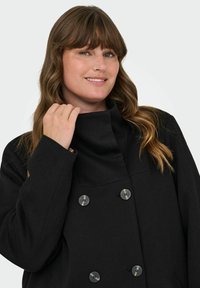 Černý svrchní oděv s vysokým límcem, dvouřadým zapínáním a kulatými knoflíky, vyrobený z hladké texturované látky.