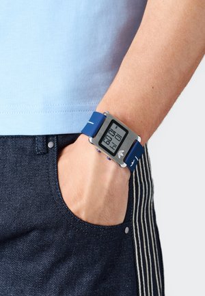 Person med högra handen i fickan på mörkblå randiga byxor, bär en blå digital klocka som visar 22:14 på handleden.