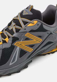 Chaussure de trail running New Balance grise et noire avec des accents jaunes, empeigne en mesh et semelle robuste pour une utilisation en extérieur.