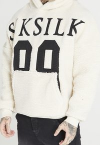 Hellbeiger Fleece-Kapuzenpullover mit schwarzer "SKSILK"-Beschriftung und "00" auf der Vorderseite, ausgestattet mit einer Kängurutasche vorn und gerippten Bündchen.