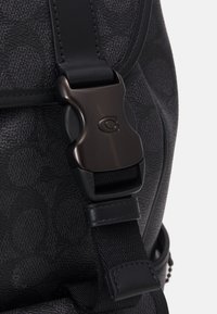 Zwarte getextureerde rugzak met een zwarte plastic gesp, een strakke schouderband en een gladde afwerking. Detail toont de hardware en textuur van het materiaal.