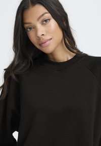 Melns krekls ar ribinātu apkakli un raglanu piedurknēm, izgatavots no mīksta auduma, ar vienkāršu dizainu un bez redzamām zīmēm.