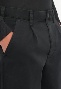 Mustadest puuvillase segu materjalist pükstest, millel on sile tekstuur, nähtavad ees olevad pleid, taskud ja kontrastne lilla tikandi detail.