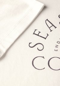 Camiseta blanca de algodón con un logo impreso en gris oscuro. La tela tiene una textura de canalé fina y cuenta con un manga plegada.