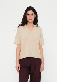 Μπεζ πλεκτό πουλόβερ με κοντά μανίκια και V-λαιμόκοψη, ελαφρώς φαρδιάς εφαρμογής με υφασμάτινο φινίρισμα, φορεμένο πάνω από σκούρο βουργουνδίσιο παντελόνι.