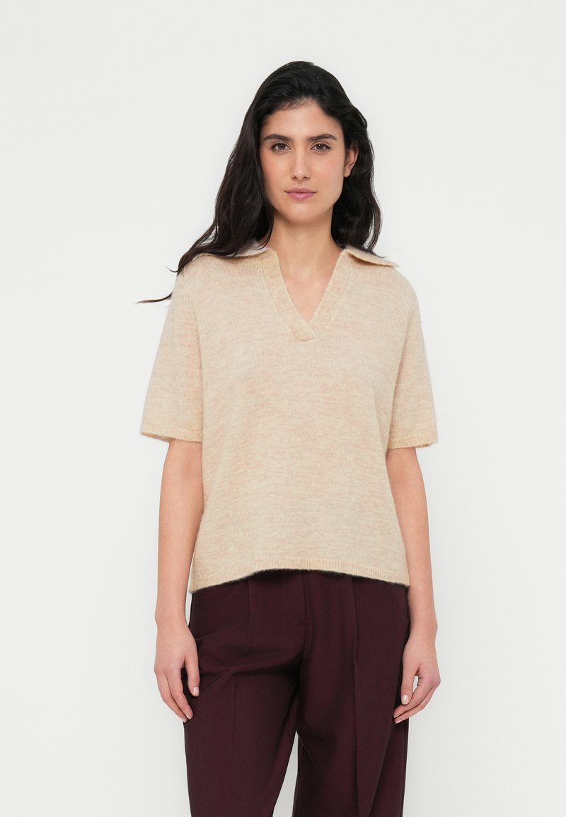 Μπεζ πλεκτό πουλόβερ με κοντά μανίκια και V-λαιμόκοψη, ελαφρώς φαρδιάς εφαρμογής με υφασμάτινο φινίρισμα, φορεμένο πάνω από σκούρο βουργουνδίσιο παντελόνι.