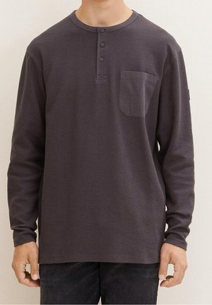 Homme portant un t-shirt henley gris foncé à manches longues avec trois boutons et une poche poitrine, debout devant un fond uni.