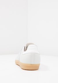 Zapatilla blanca con parte superior de cuero liso, detalles texturizados y suela de goma marrón claro; cuenta con un talón acolchado y diseño a rayas en los lados.
