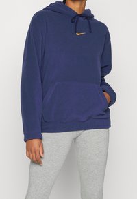Mörkblå huvtröja i mjukt material, med en framsidans ficka och en liten gyllene Nike-logga på bröstet. Ljust grå leggings.