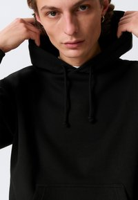 Must kapuutsiga dressipluus kähriktaskuga, millel on nöörid ja pehme tekstuur. Minimalistlik disain, lõdva istuvusega ja ilma nähtavate mustriteta.