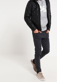 Chaqueta negra acolchada sobre una sudadera gris con logo blanco, jeans oscuros y zapatillas marrón claro. Diseño simple, casual y funcional.