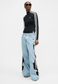 Άτομο που φοράει μαύρο τοπ Adidas με λευκές ρίγες και σκισμένο ανοιχτό μπλε τζιν παντελόνι με φαρδιά μπατζάκια, με μαύρα υφασμάτινα πάνελ από κάτω.