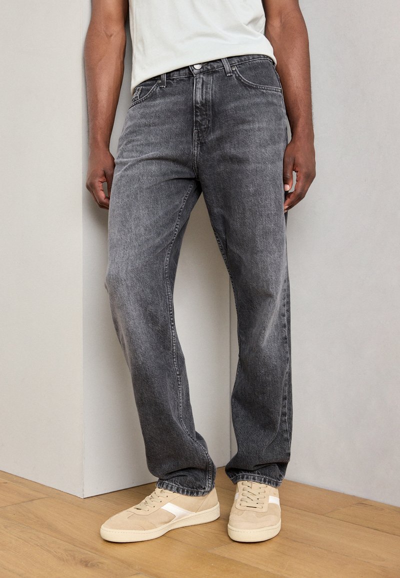 Jeans de mezclilla gris con un corte holgado, ligero desgaste y diseño clásico de cinco bolsillos, combinados con zapatillas beige claras.