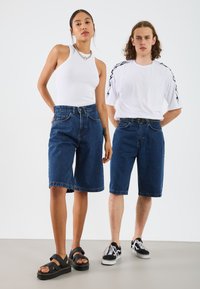 Två personer står sida vid sida, klädda i jeansshorts och avslappnade toppar. Kvinnan har på sig en vit ribbad linne och mörkblå shorts. Mannen bär en vit skjorta med svarta accenter och matchande blå shorts. Båda har på sig avslappnade skor.