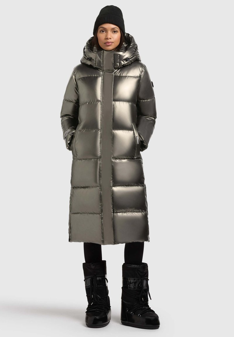 Lange, puffy parka in metallic olijfgroen met hoge kraag, capuchon en ritssluiting aan de voorkant. Gecombineerd met zwarte, met bont gevoerde winterlaarzen.
