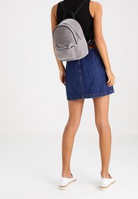 Mochila de canalé gris con dos compartimentos con cremallera y correas ajustables, llevada sobre una falda de mezclilla y alpargatas blancas.