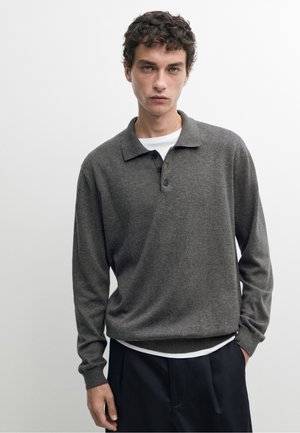 Junger Mann mit lockigem Haar, der einen dunkelgrauen Langarm-Polo-Pullover über einem weißen Hemd und schwarzen Hosen trägt, steht vor einem schlichten Hintergrund.
