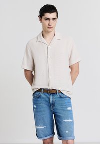 Camicetta corta beige chiaro con tessuto texture e chiusura a bottoni, abbinata a pantaloni corti in denim azzurro chiaro con dettagli sfrangiati.