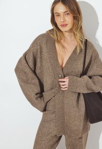 Brūns vilnas kardigāns ar pārspīlētu piegriezumu, dziļu V veida kaklu, ribotiem piedurkņu galiem un vienu pogu. Stilizēts ar atbilstošām biksēm un melnu somu.