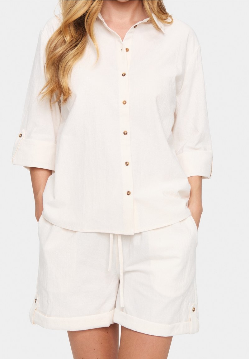 Camicia bianca di lino con bottoni davanti e maniche arrotolate, abbinata a pantaloni corti coordinati con vita elastica e orli arrotolati.