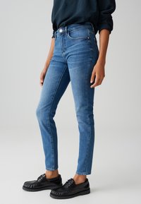 Personne portant un jean slim bleu taille mi-haute et des mocassins noirs à lacets sur un fond gris uni.