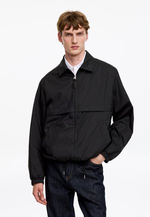 Ung mand iført en sort lynlåstrøje over en hvid skjorte og mørke jeans, der står med den ene hånd i lommen mod en hvid baggrund.
